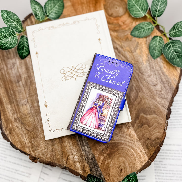 The front cover of a phone case inspired by Beauty and the Beast by Gabrielle-Suzanne de Villeneuve on a white book, on a slice of wood, with greenery behind it.
