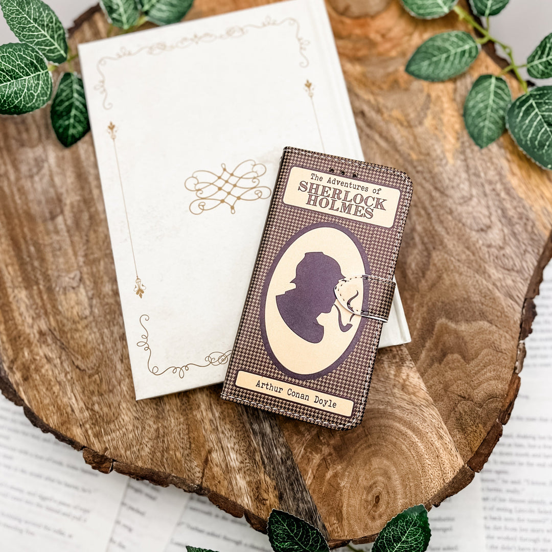 The front cover of a book-shaped phone case inspired by The Adventures of Sherlock Holmes by Sir Arthur Conan Doyle. The phone case sits on a white book cover on a wood slice surrounded by greenery.