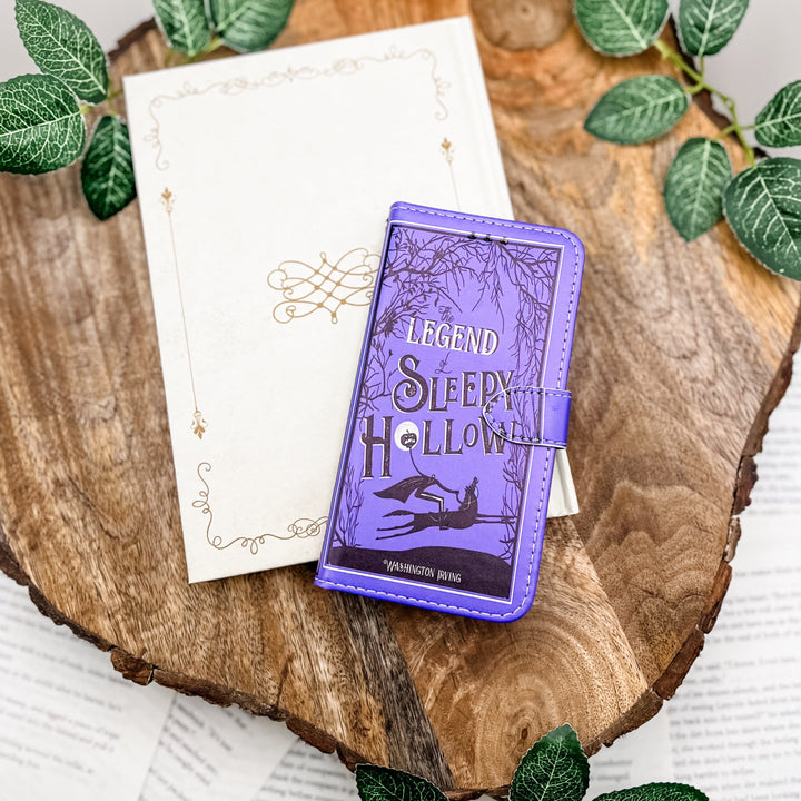 The front of a book-shaped phone case inspired by The Legend of Sleepy Hollow by Washington Irving. The phone case sits on a white book cover on top of a wood slice surrounded by greenery.