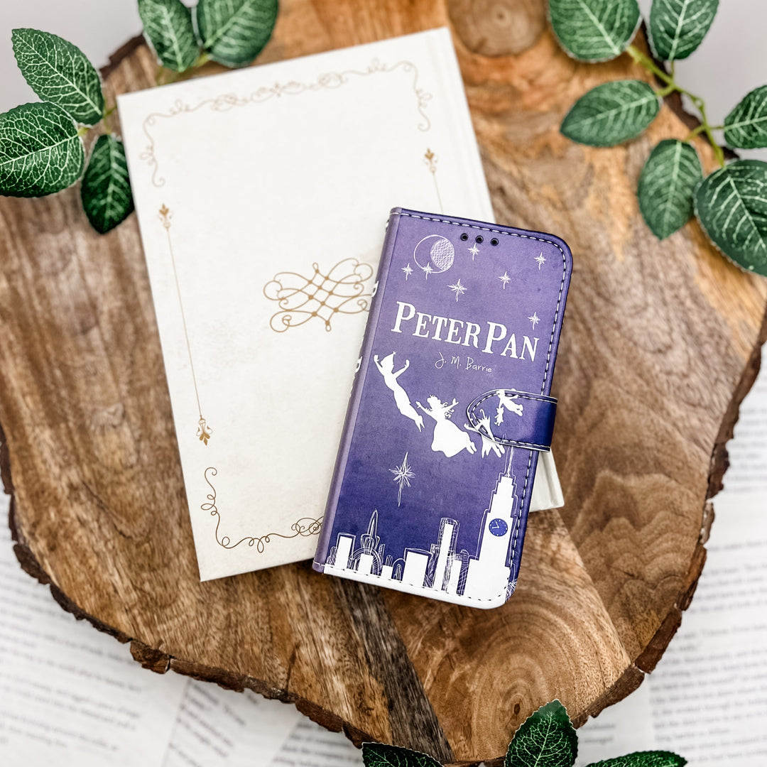 The front cover of a book-shaped phone case inspired by Peter Pan by J.M. Barrie. The phone case is on top of a white book cover on top of a wood slice surrounded by greenery.