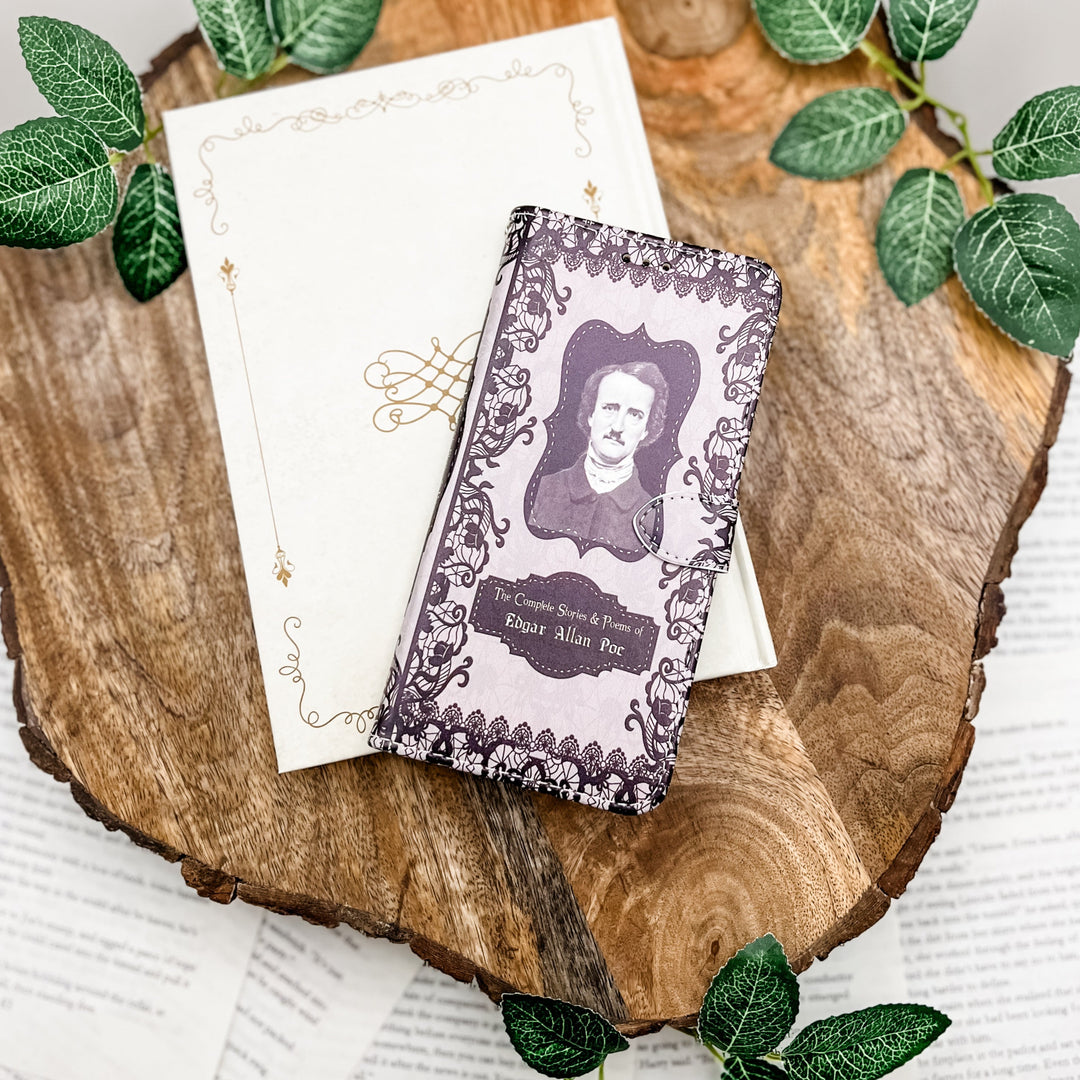 The front cover of a phone case inspired by The Complete Stories & Poems of Edgar Allan Poe. The phone case sits on a white phone case on a wood slice on a white background. Greenery surrounds it.