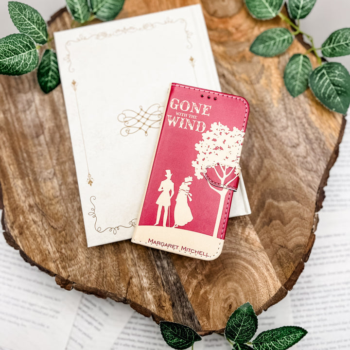 The front of a phone case inspired by Gone With the Wind by Margaret Mitchell. The phone case sits on a white book cover on a wood slab surrounded by greenery.