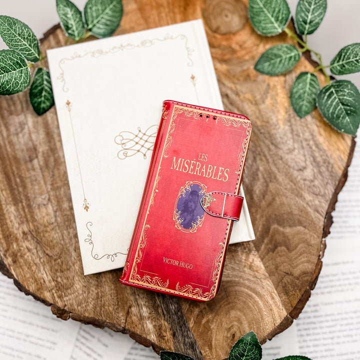 The front of a book shaped phone case inspired by Les Miserables by Victor Hugo. The phone case sits on a white book on a wood slice surrounded by greenery.
