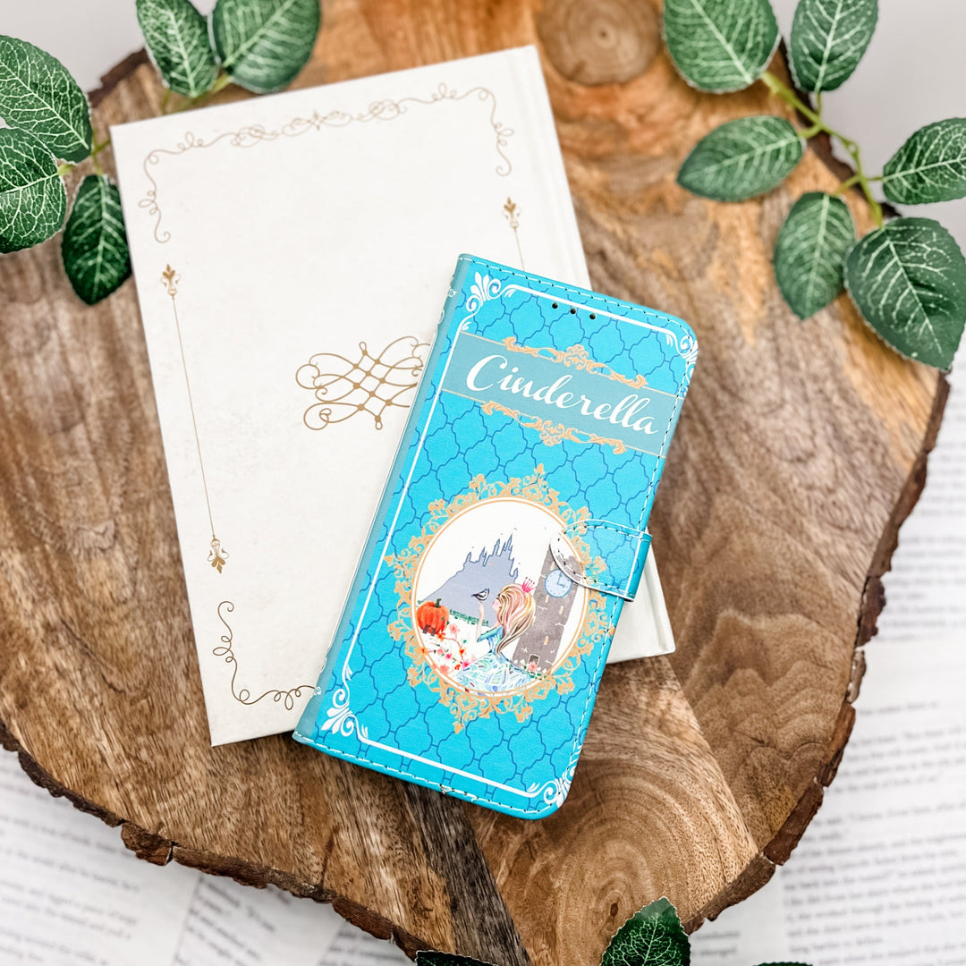 The front cover of a phone case inspired by Cinderella. The phone case sits on a closed white book on a wood slice surrounded by greenery.