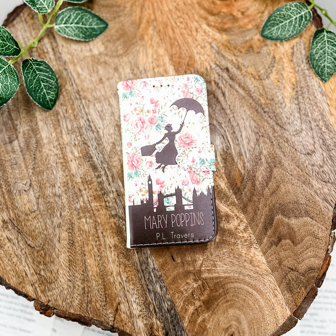 The front cover of a book-shaped phone case inspired by Mary Poppins by P.L. Travers. The phone case sits on a wood slice surrounded by greenery.