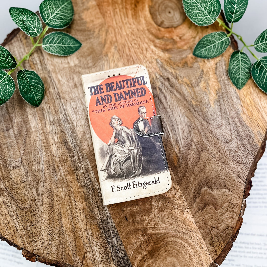 The front cover of a phone case featuring The Beautiful and Damned by F. Scott Fitzgerald. The phone case sits on a wood slice surrounded by greenery.