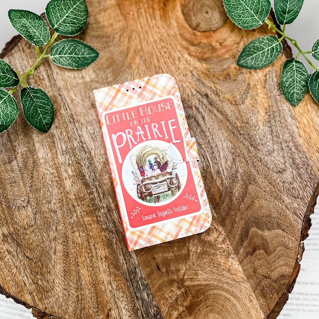 The front of a book shaped phone case inspired by Little House on the Prairie by Laura Ingalls Wilder. The phone case sits on a wood slice surrounded by greenery.