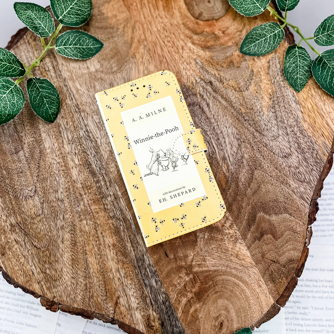 The front cover of a book-shaped phone case inspired by Winnie-the-Pooh by A.A. Milne. The phone case sits on a wood slice surrounded by greenery.