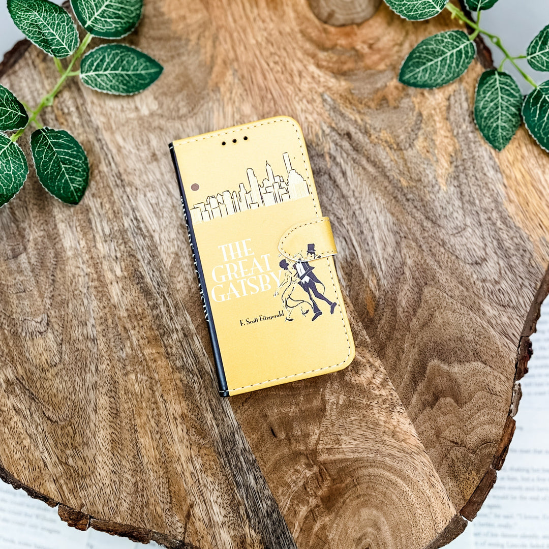 The front of a book-shaped phone case inspired by The Great Gatsby by F. Scott Fitzgerald. The phone case sits on top of a wood slice and surrounded by greenery.