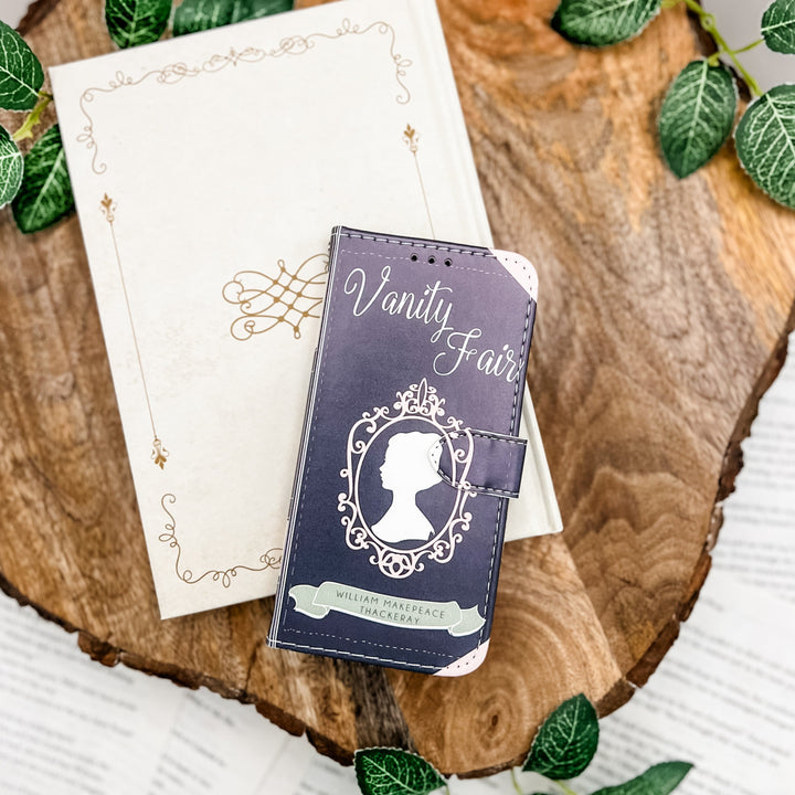 The front cover of a book-shaped phone case inspired by Vanity Fair by William Makepeace Thackeray. The phone case sits on a white book cover on a wood slice surrounded by greenery.