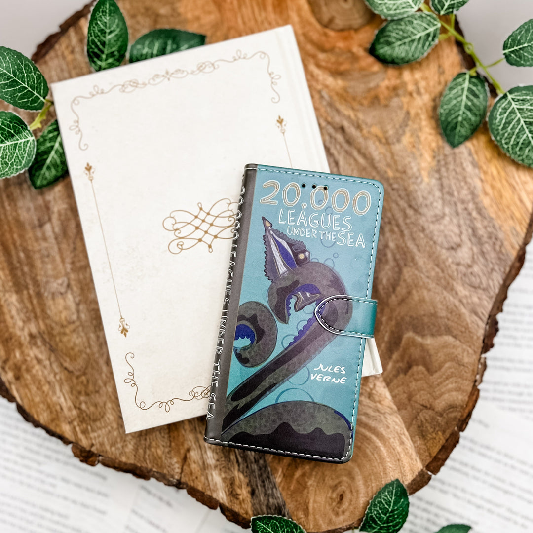 The front cover of a phone case inspired by 20,000 Leagues Under the Sea by Jules Verne. The phone case sits on a white book on a wood slice surrounded by greenery.