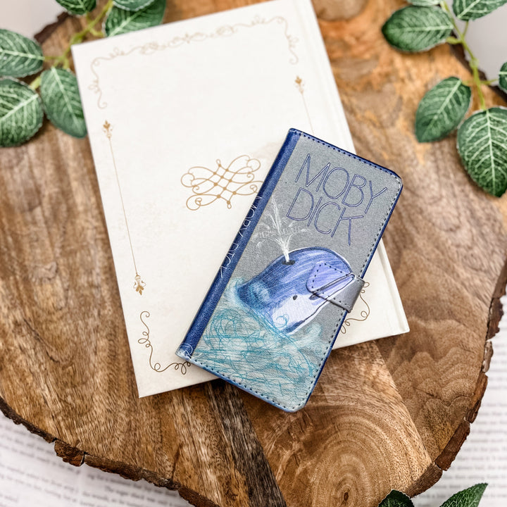 The front of a book-shaped phone case inspired by Moby Dick by Herman Melville. The phone case sits on a white book cover on a wood slice surrounded by greenery.