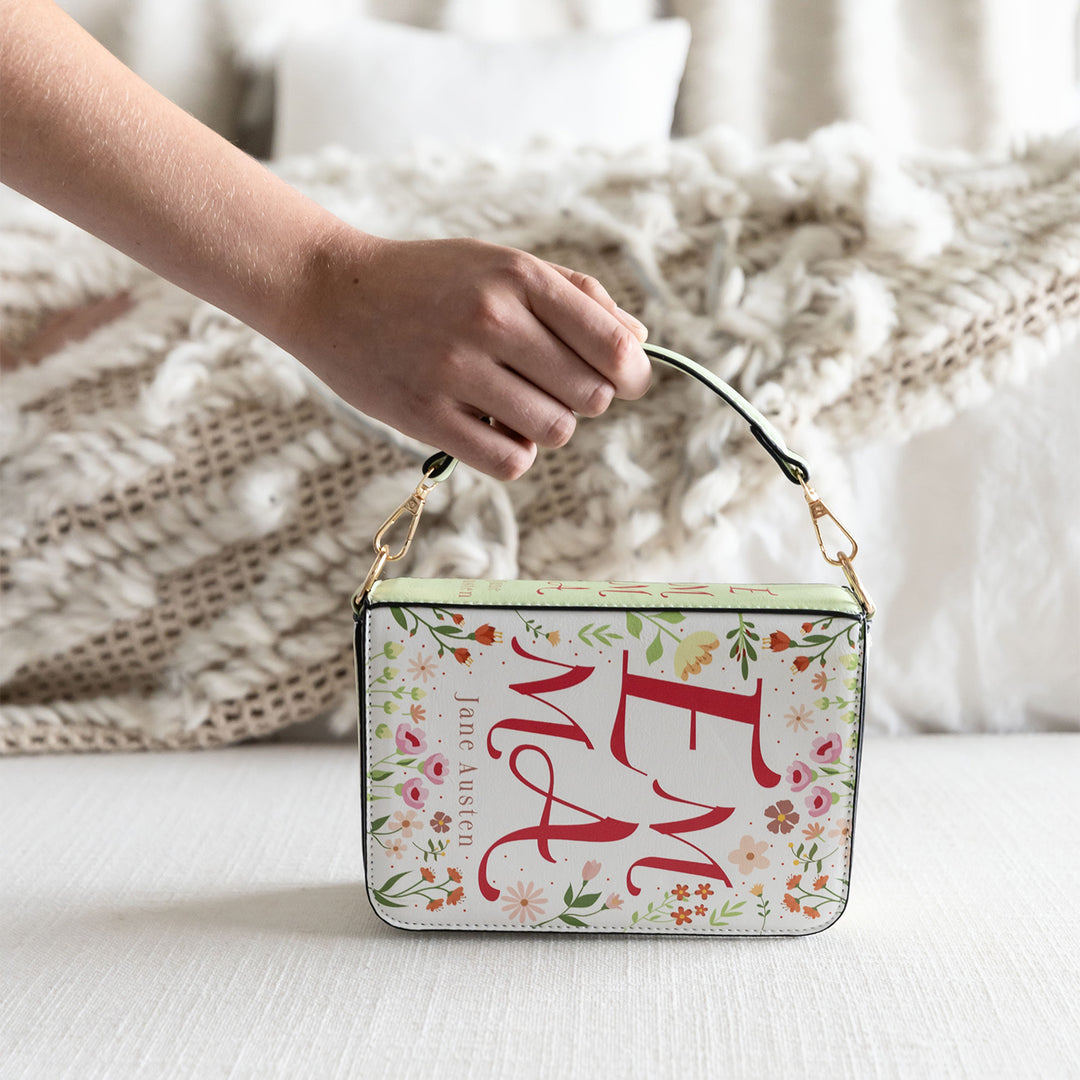 A book-shaped purse inspired by Emma by Jane Austen. A white hand grips the short handle of the purse. The purse stands on its side with a white, cozy background.