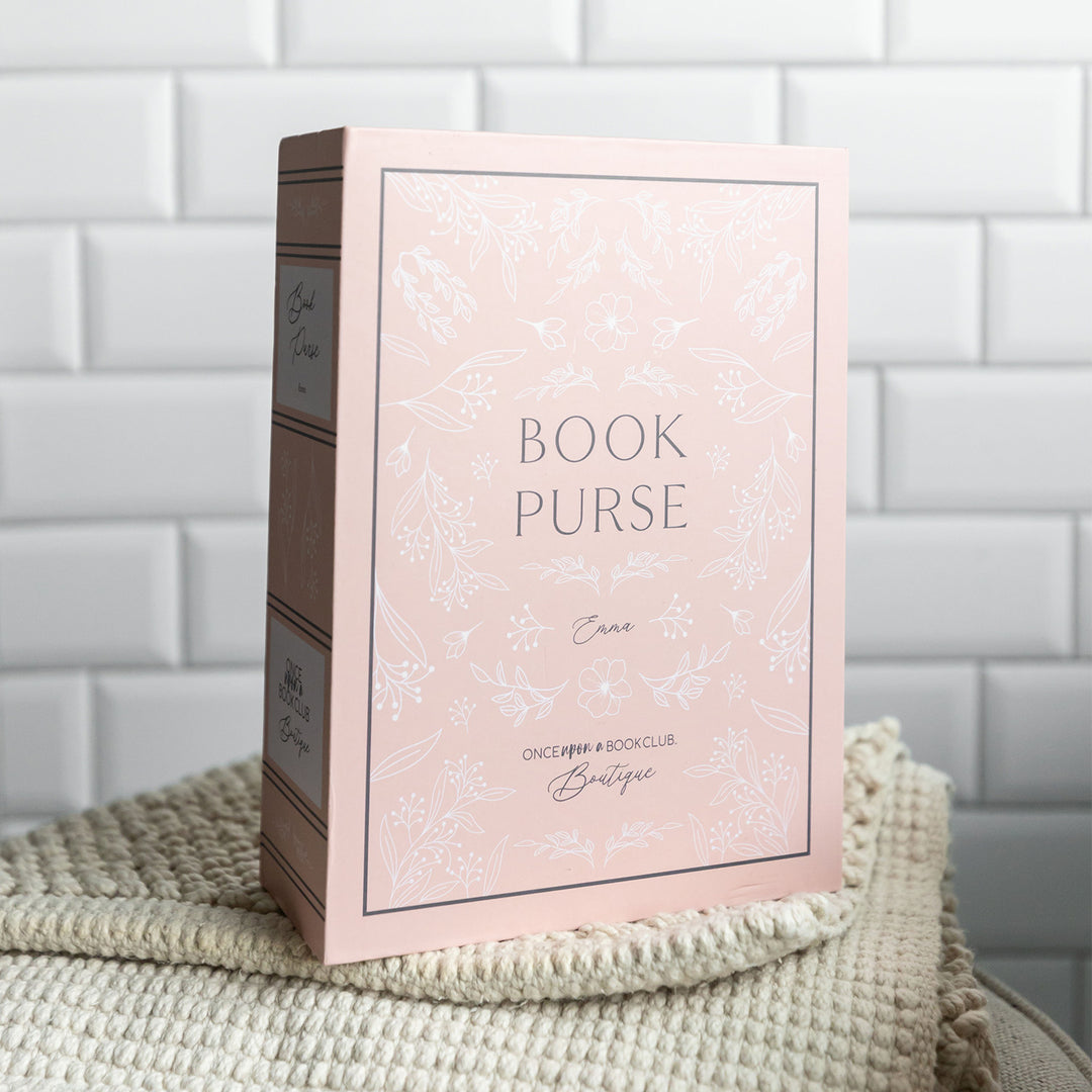 The book-shaped box used to package our book purse. The box is light pink and features the words "Book Purse" "Emma" "Once Upon a Book Club Boutique" on the front cover and spine with floral detailing surrounding it.