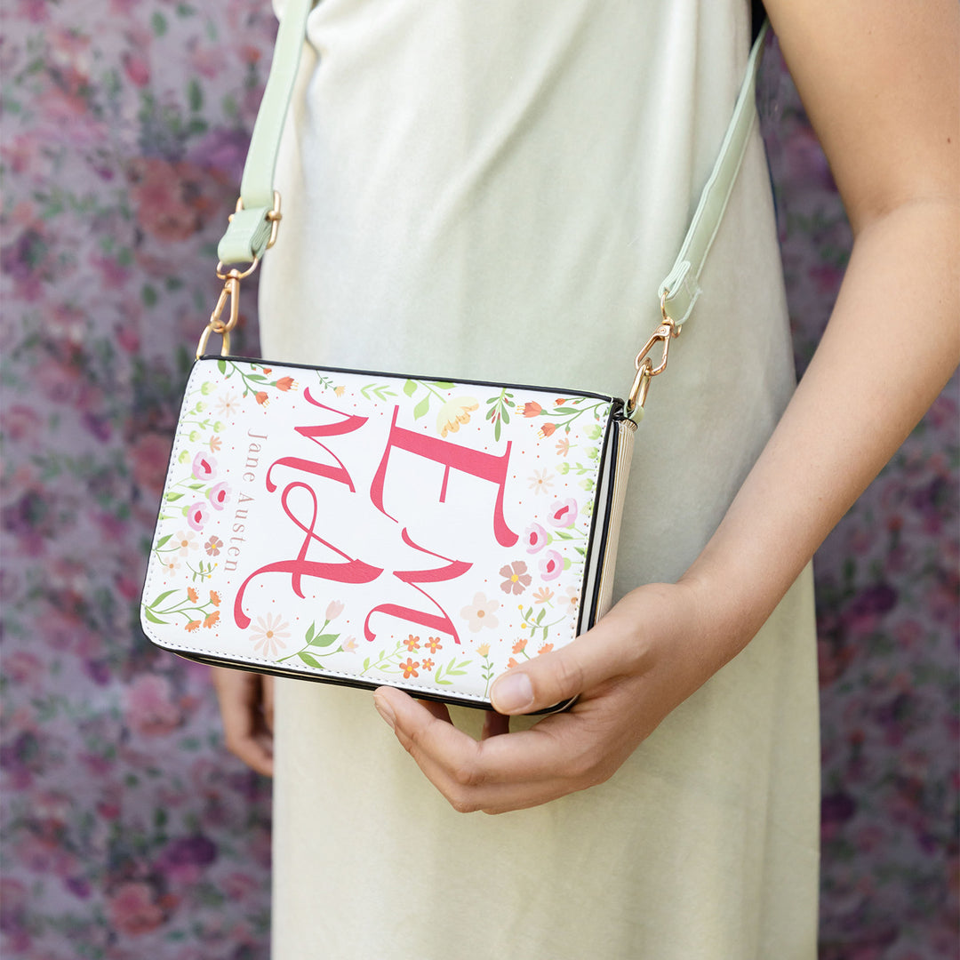 A book-shaped purse inspired by Emma by Jane Austen. A white woman holds the purse against her green dress. The crossbody strap is utilized to showcase it pairs with an outfit.