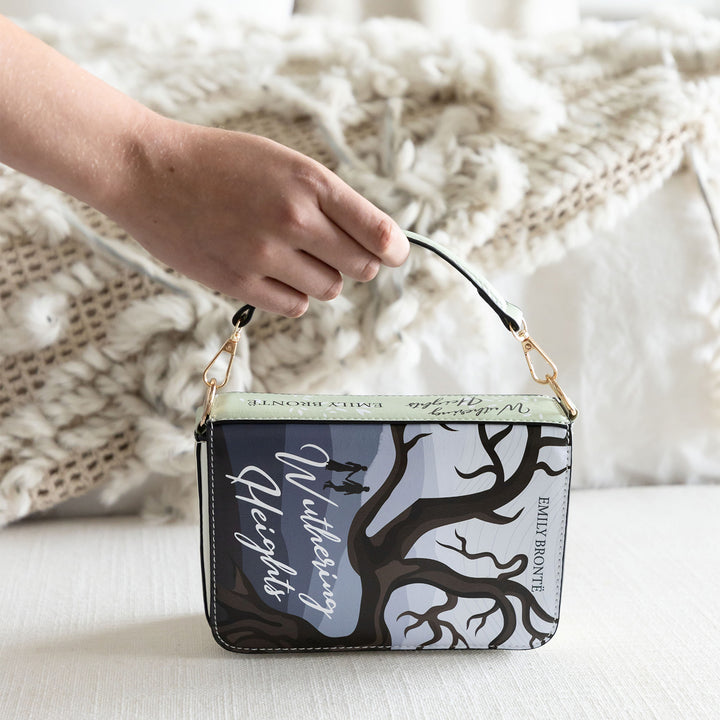 A book-shaped purse inspired by Wuthering Heights by Emily Brontë. A white hand grips the short handle of the purse and the spine is visible beneath the handle. The front cover is facing the camera.