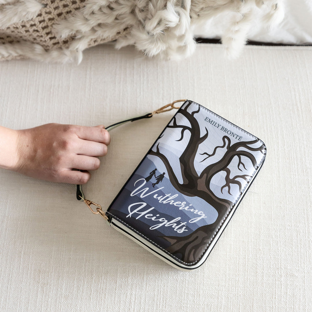 A book-shaped purse inspired by Wuthering Heights by Emily Brontë. A white hand grasps the short handle to show its length. The front cover of the purse is visible on a cream-toned, fabric background.