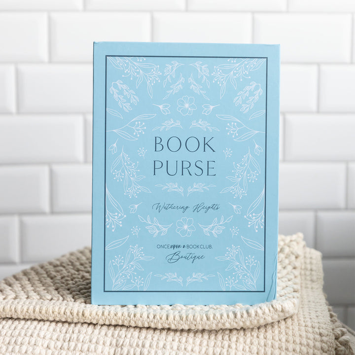 The book-shaped box used to package our book purse. The box is light blue and features the words "Book Purse" "Wuthering Heights" "Once Upon a Book Club Boutique" on the front cover and spine with floral detailing surrounding it.