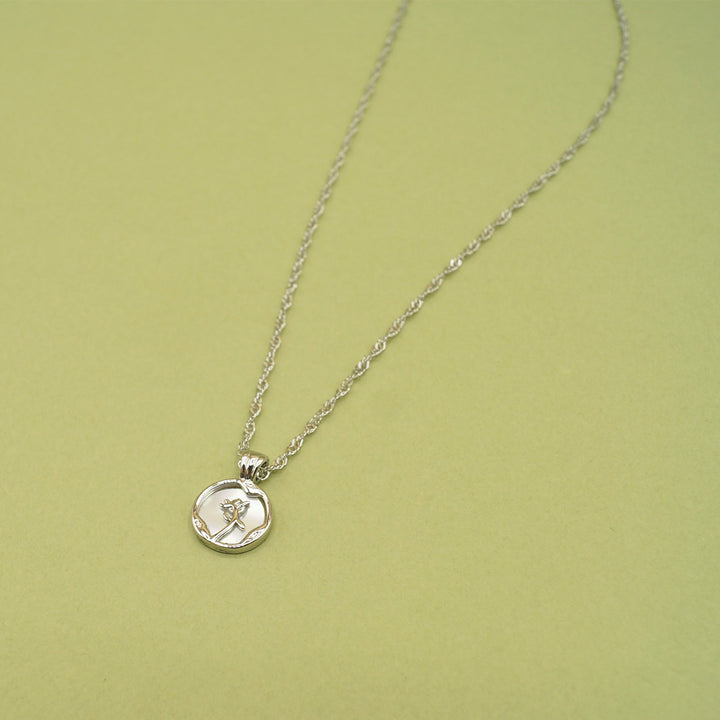 The silver pendant featuring a small disc with a rose shape in the middle of it sits on green background.