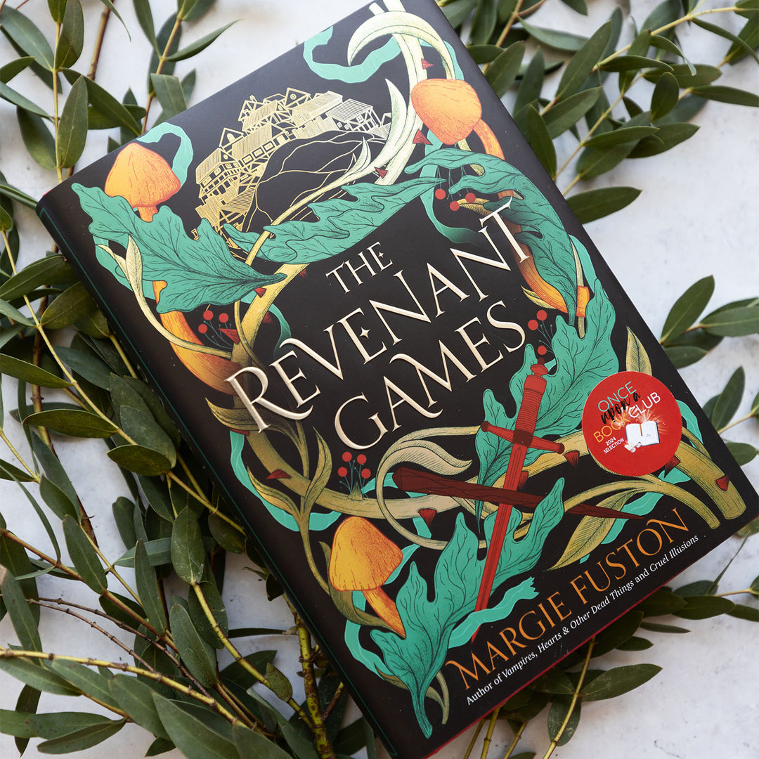 A hardcover edition of The Revenant Games by Margie Fuston sits on greenery on a white background.