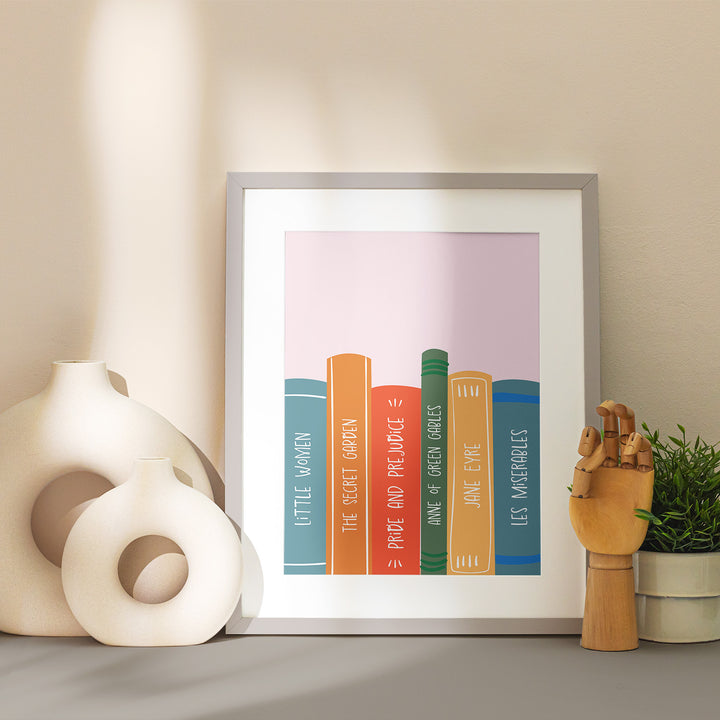 A grey matted frame sits on a table showcasing the "Book Spines" art print. Around the frame is stylized with vases and home decor in neutral tones.