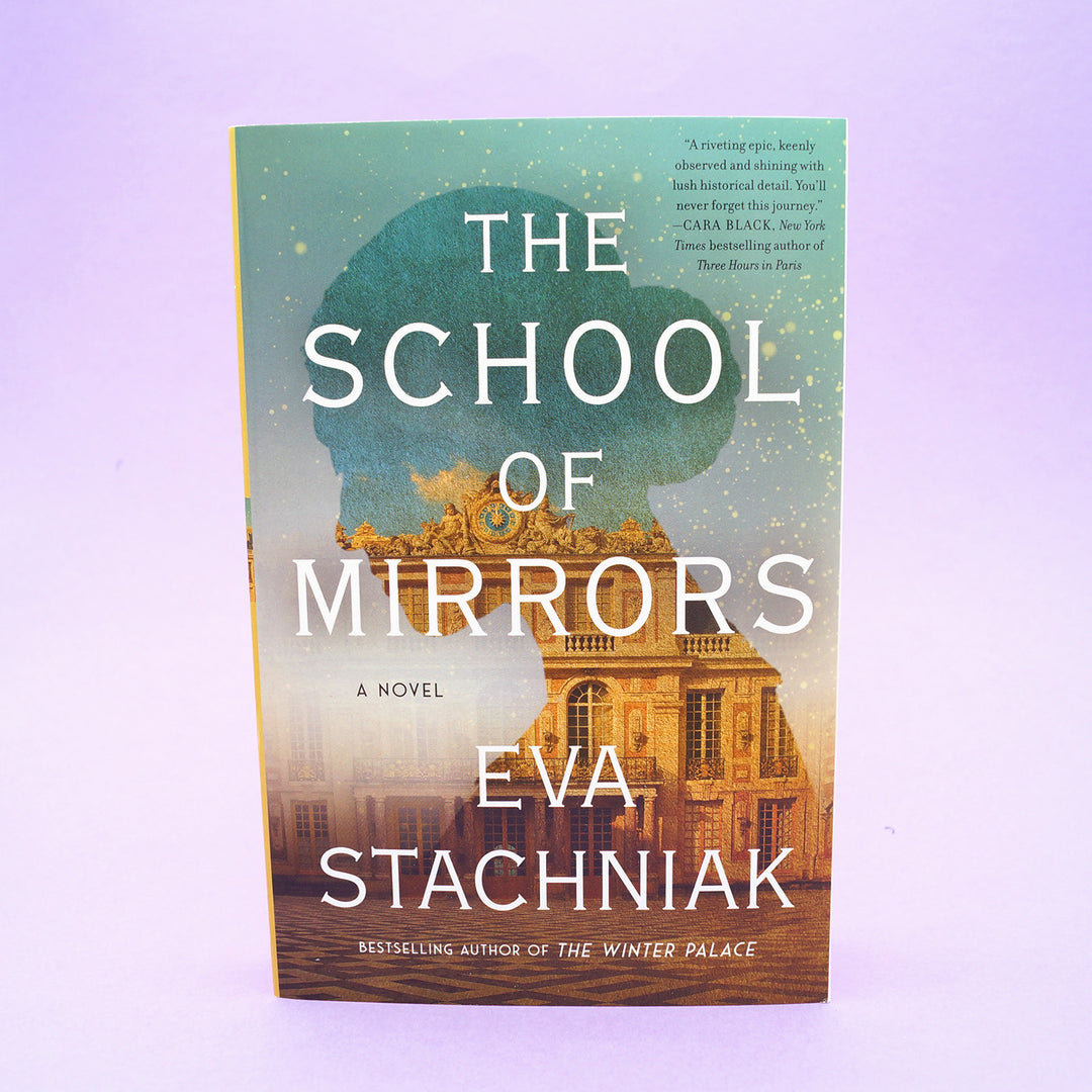 A paperback copy of The School of Mirrors by Eva Stachniak sits on a purple background.