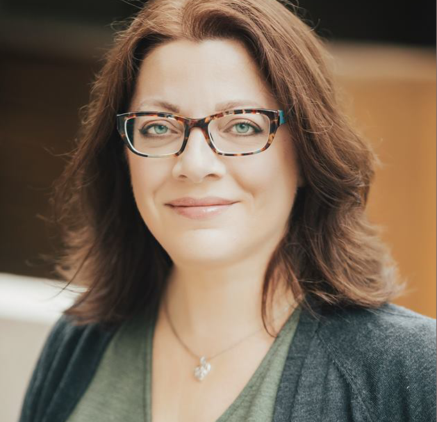 Author Simone St. James smiling at the camera. She is a middle-aged white woman with reddish-brown hair, tortoiseshell glasses, and green eyes. She is wearing a green and grey top and a pendant necklace.