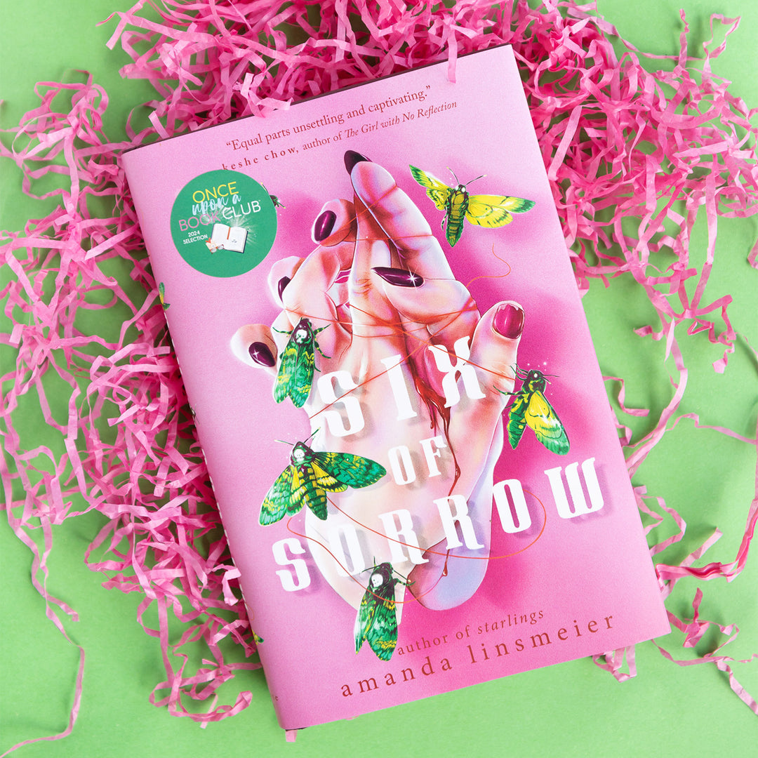 a hardcover copy of Six of Sorrow by Amanda Linsmeier on pink shredded paper and a green background.