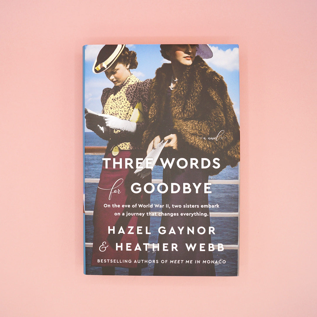 A hardcover edition of Three Words for Goodbye by Hazel Gaynor and Heather Webb sits on a pink background.