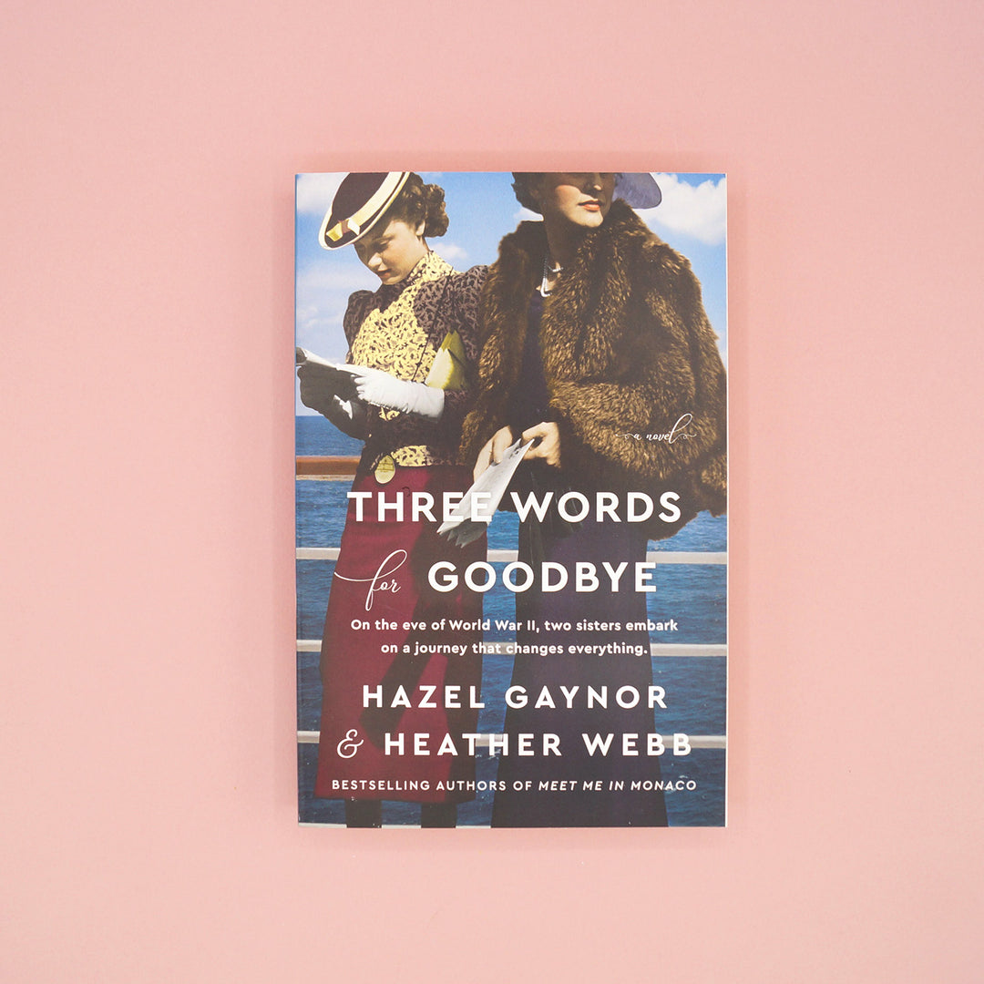 A paperback edition of Three Words for Goodbye by Hazel Gaynor and Heather Webb sits on a pink background.