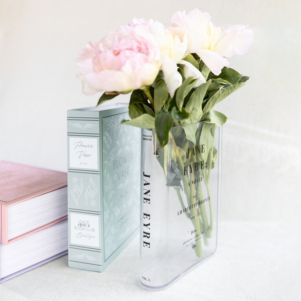 A clear acrylic book-shaped vase printed to look like a copy of Jane Eyre by Charlotte Bronte sits on a white background. The vase contains a small bouquet of pink and white flowers. The green book-shaped box stands next to it.