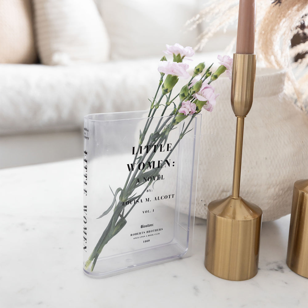 A clear acrylic book-shaped vase printed to look like a copy of Little Women by Louisa M. Alcott sits on a white surface. The vase holds a small bouquet of pink flowers. Next to the vase is neutral-toned home decor.