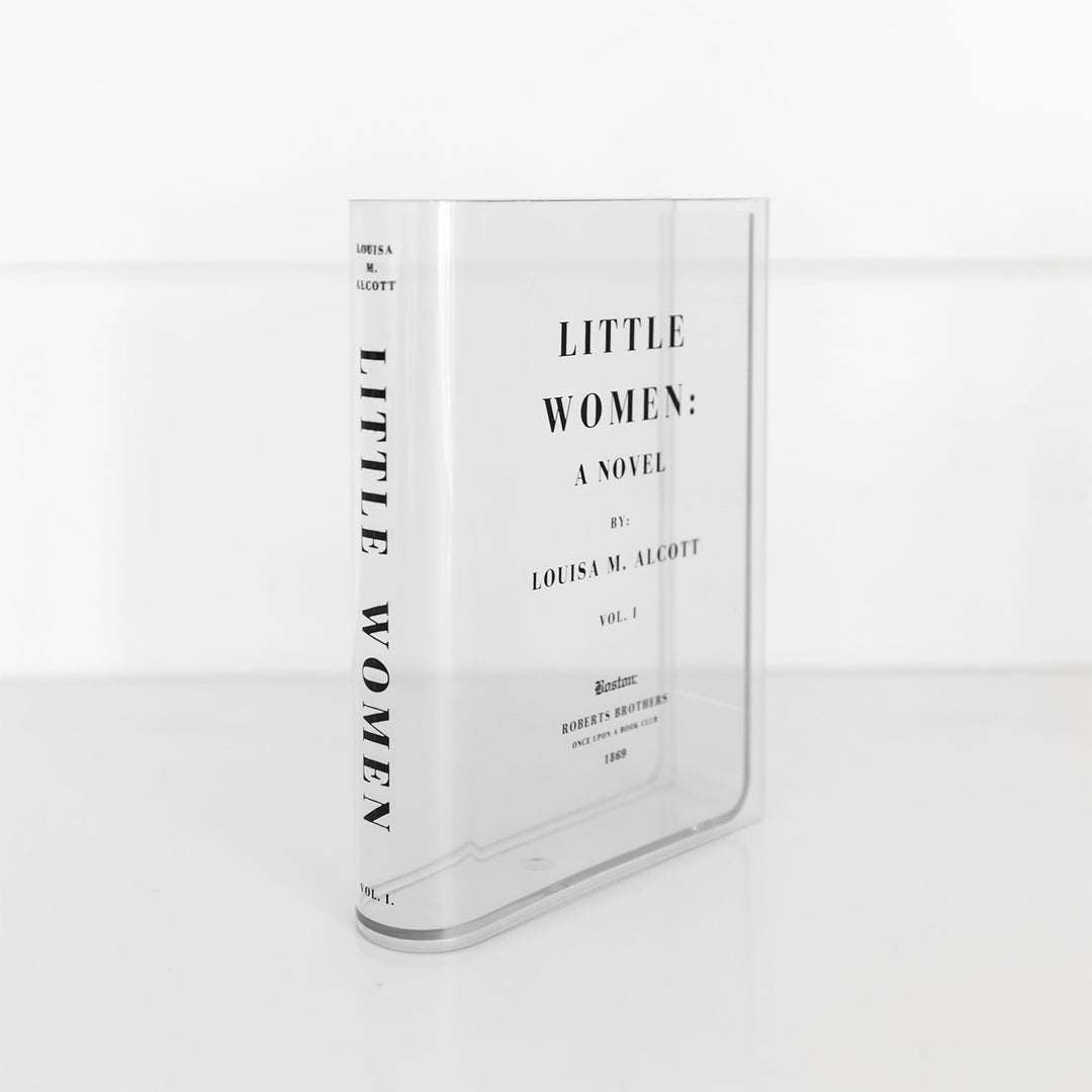 A clear acrylic book-shaped vase printed to look like a copy of Little Women by Louisa M. Alcott sits on a white surface. The spine of the 'book' faces outward and can be read in black bold font.