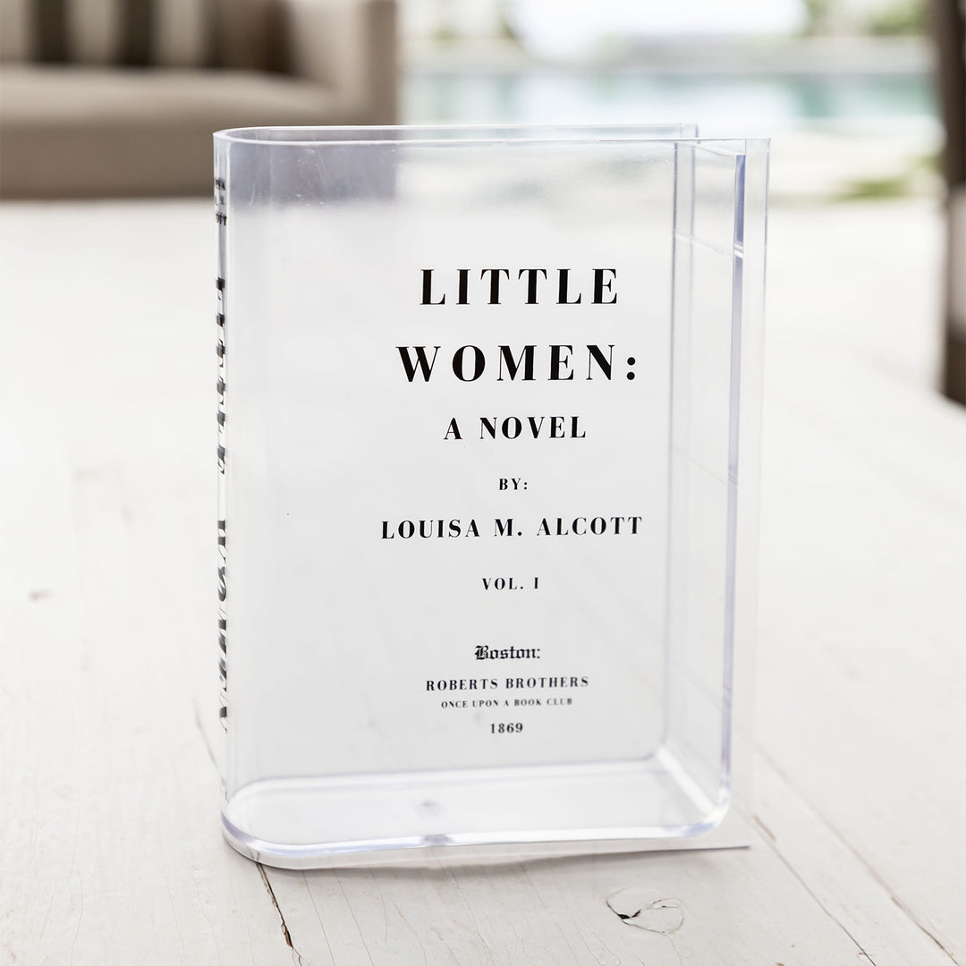 A clear acrylic book-shaped vase printed to look like a copy of Little Women by Louisa M. Alcott sits outdoors.