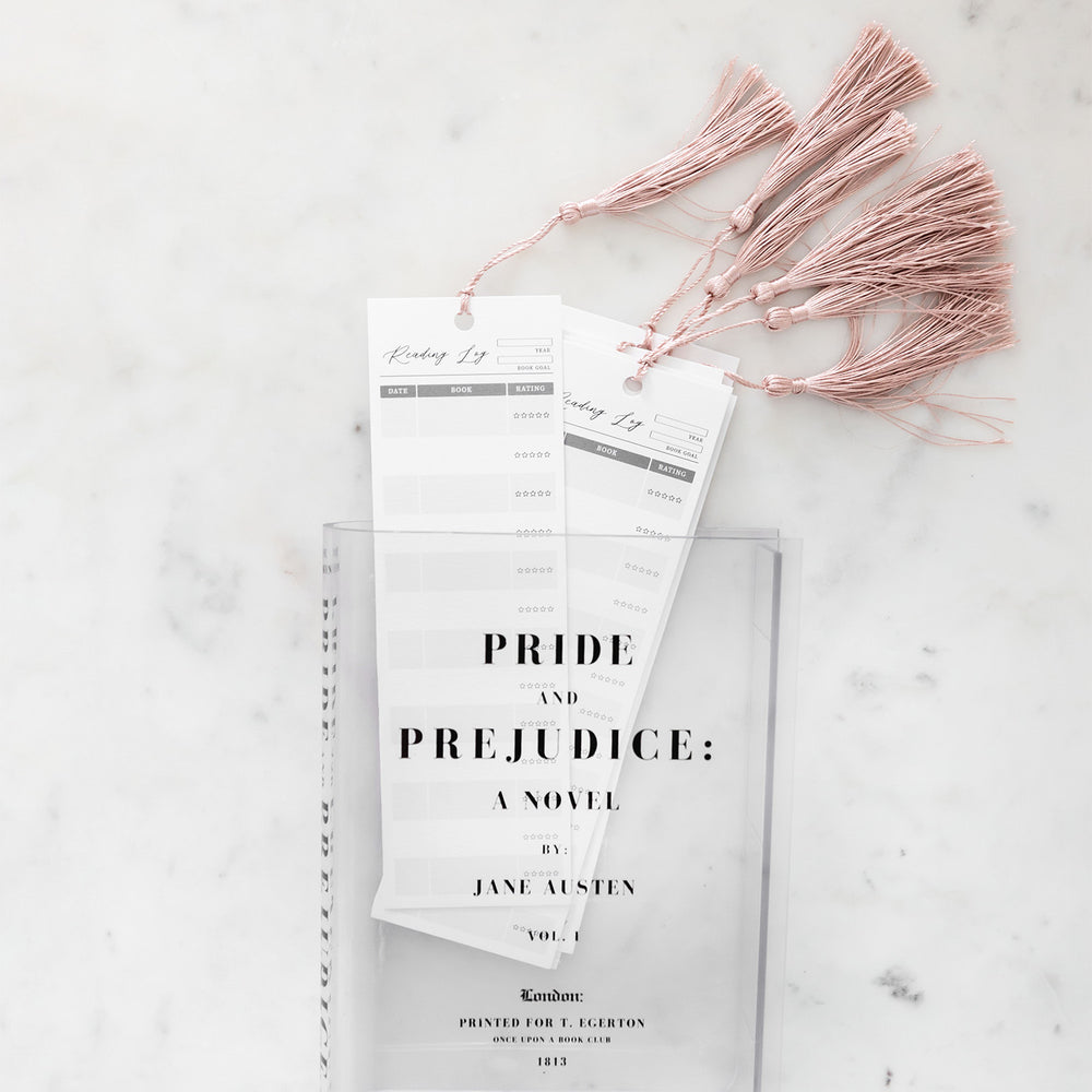 A clear acrylic book-shaped vase printed to look like a copy of Pride and Prejudice by Jane Austen lays on a white marbled surface. Inside the vase is a collection of white reading log bookmarks with pink tassels.