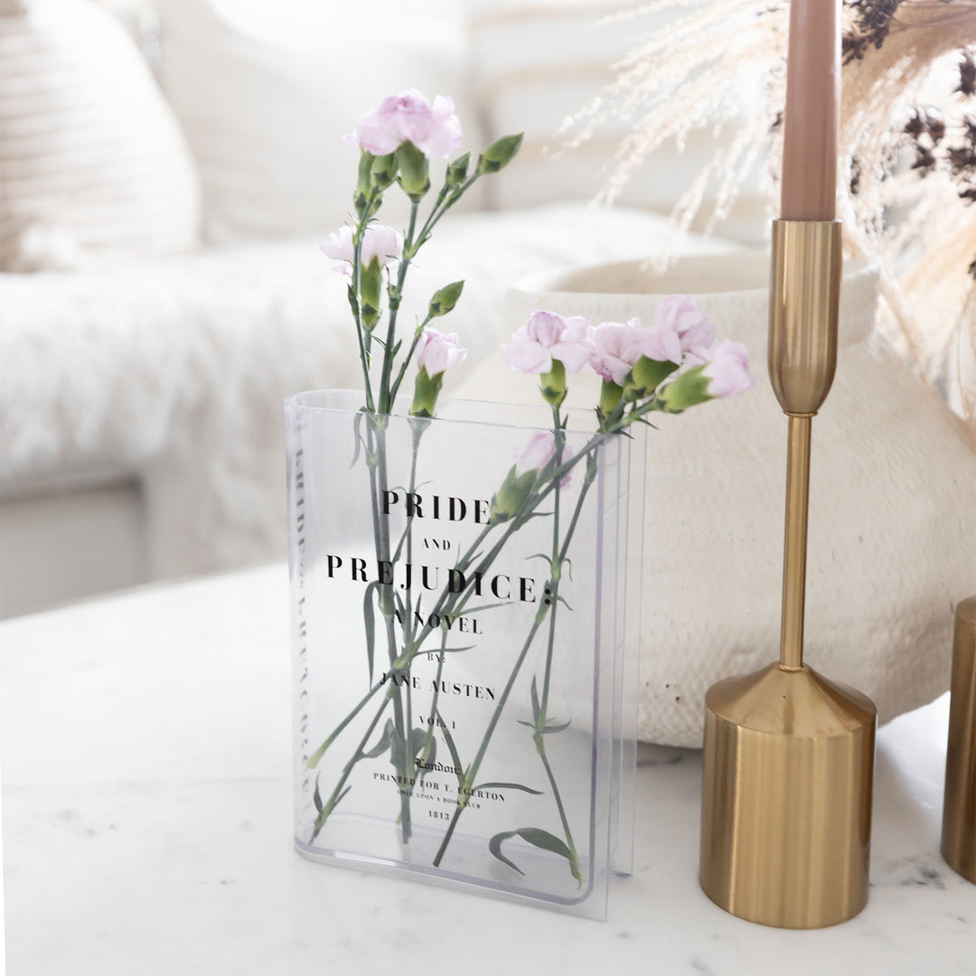 A clear acrylic book-shaped vase printed to look like a copy of Pride and Prejudice by Jane Austen sits on a white surface. Inside the vase is a small arrangement of pink and white flowers. Next to the vase is an array of neutral toned home decor.