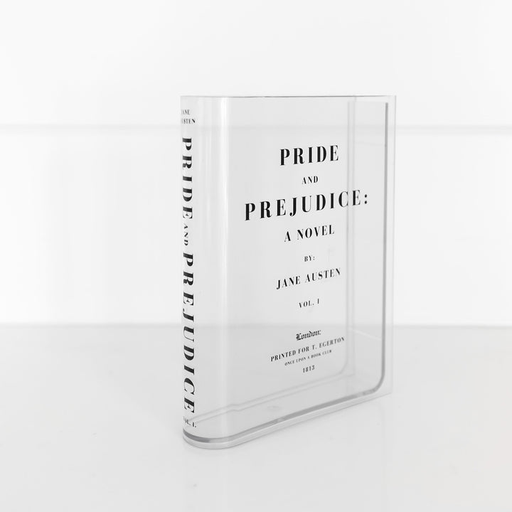 A clear acrylic book-shaped vase printed to look like a copy of Pride and Prejudice by Jane Austen sits on a white surface. The spine of the vase is angled toward the camera and the book title on the spine is visible.