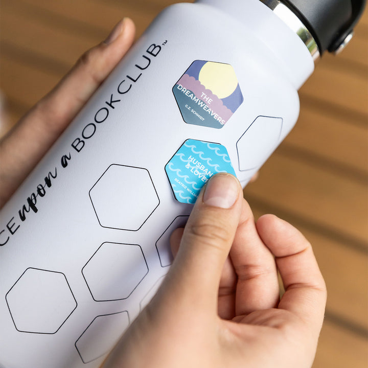 The white water bottle is held horizontally by a pair of white hands. A blue hexagon sticker showing the book title "Husbands and Lovers" with a wave pattern is being placed into an empty hexagon on the side of the bottle. Above it, a sticker with a moon and purple and teal detailing reads "The Dreamweavers" and is already stuck.