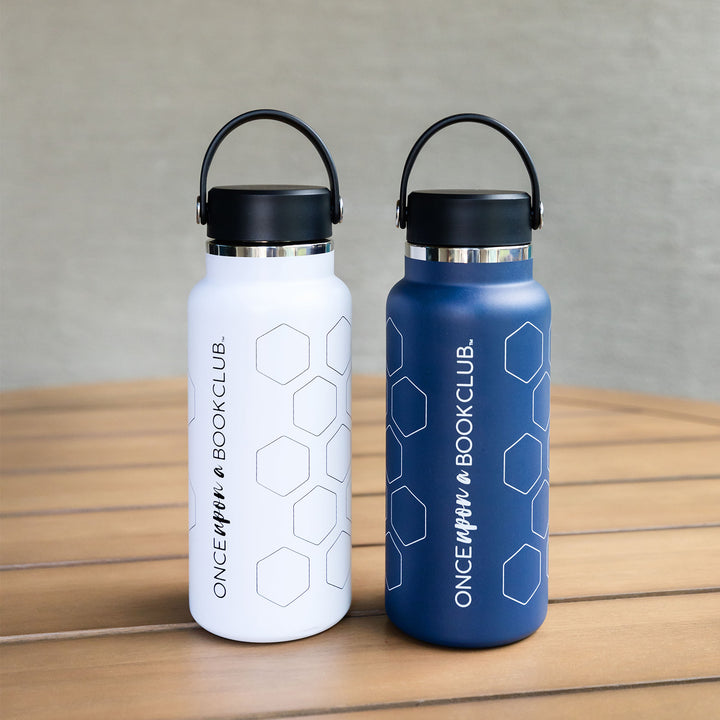 Two water bottles sit on a wooden table. One water bottle is white with a black lid and has black writing on the metal exterior that reads "Once Upon a Book Club" with empty hexagons wrapping around the bottle. The other is dark blue with a black lid and has white writing on the metal exterior that reads "Once Upon a Book Club" with empty hexagons wrapped around the bottle.