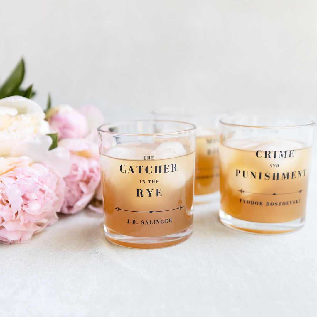 A set of whiskey glasses inspired by classic book titles. Visible are The Catcher in the Rye by J.D. Salinger and Crime and Punishment by Fyodor Dostoevsky. A third glass is visible in the background. Each glass is filled with an amber-colored liquid. Pink flowers lay on the white background to the left of the glasses.