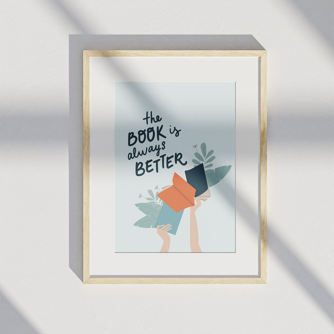 A matted wooden frame hangs on a white wall and displays the "The Book Is Better" art print. A shadow from a window is visible over the art print.