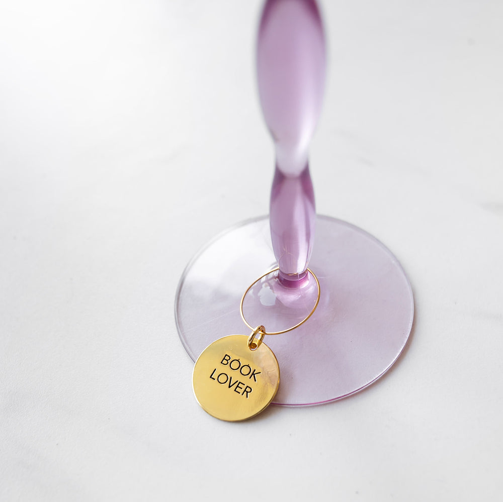 A gold "Book Lover" wine charm is wrapped around the stem of a purple wine glass.