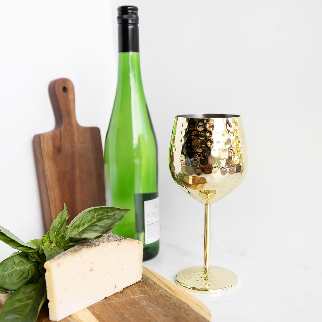 A gold wine glass stands next to an empty green bottle of wine, a wedge of cheese, and a wooden cutting board.