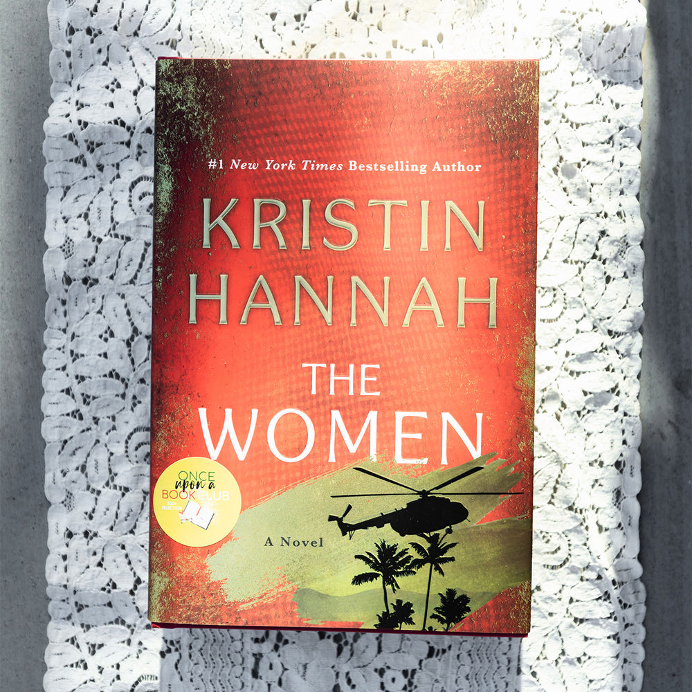 A hardcover copy of The Women by Kristin Hannah sits on a white lace background.