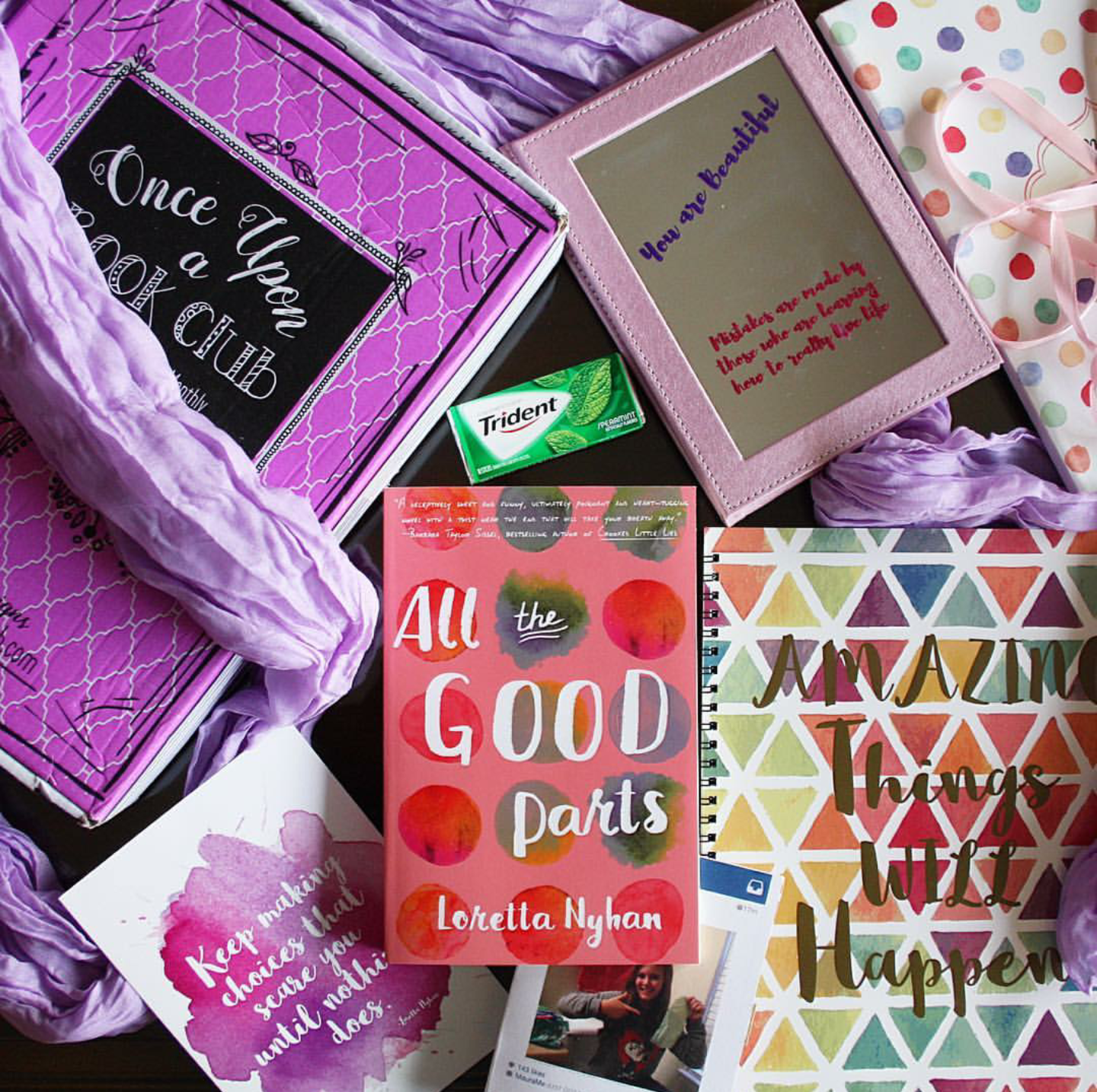 A paperback copy of All the Good Parts by Loretta Nyhan sits next to a pink Once Upon a Book Club box. Surrounding it are the unwrapped gifts, including a purple scarf, travel mirror with fabric case and motivational message on it, a rainbow notebook, a photograph, and a pack of gum.