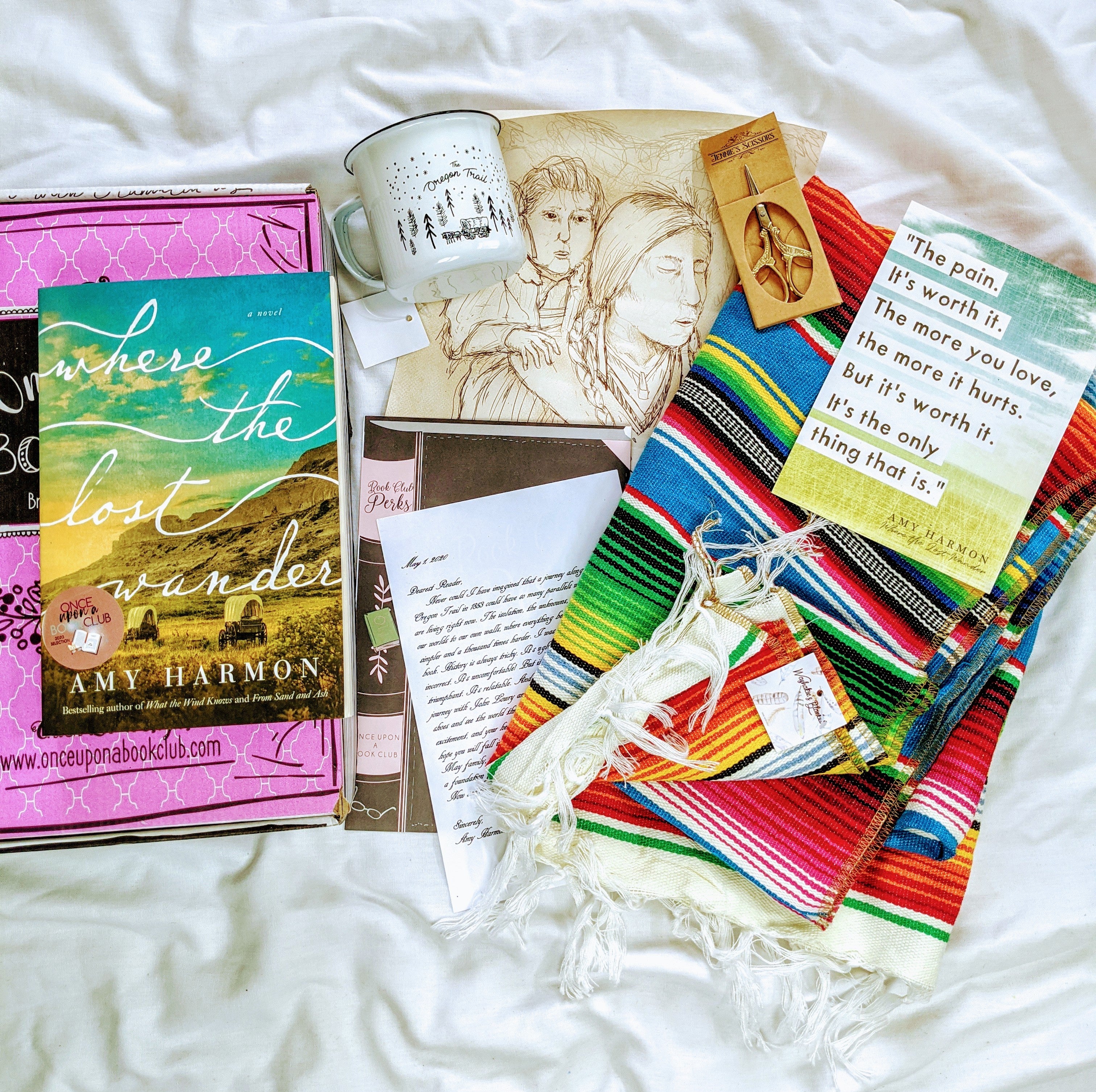 A paperback copy of Where The Lost Wander by Amy Harmon sits on top of a pink Once Upon a Book Club box. Next to this are the unwrapped gifts pulled from the story, including a multi-colored blanket, a small pair of scissors, a letter, a mug, and a drawing.